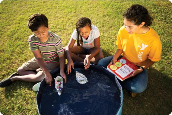 two campers and a counselor testing out their tin foil boats in a small kiddie pool